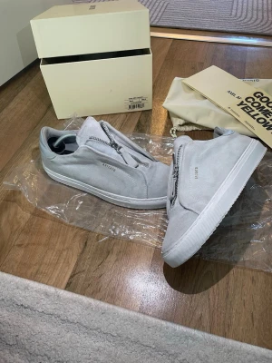 Ljusgrå sneakers med dragkedja från Axel Arigato  - Snygga ljusgrå sneakers i mocka med dragkedja framtill istället för klassiska snören. Skorna har en stilren och minimalistisk design med vit sula och diskret text på sidan. Perfekta för dig som gillar en clean och modern look. Storlek 40 passar 41 obs de är lite smutsiga 