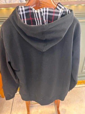 Svart hoodie med dragkedja och rutigt foder - Stilren svart hoodie från Burberry med dragkedja framtill och klassiskt rutigt foder i huvan. Hoodien har fickor på magen, ribbade muddar och en diskret logga på bröstet. Perfekt för dig som gillar en clean look med exklusiva detaljer.