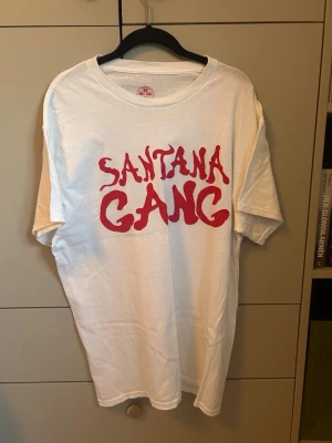 Vit Santana Gang t-shirt med tryck - Vit t-shirt med röd text 'Santana Gang' på framsidan och en stor röd örn med texten 'West' på baksidan. T-shirten har rund hals och korta ärmar. Perfekt för dig som gillar streetwear och unika prints.