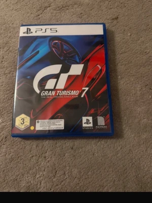 Gran Turismo 7 till PS5 - Gran Turismo 7 till PlayStation 5, ett racingspel med realistisk körkänsla. Omslaget är blått med röda och blå detaljer och spelet levereras i ett klassiskt plastfodral. Perfekt för dig som gillar bilar och tävlingar.
