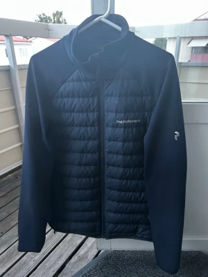 Mörkblå jacka från Peak Performance - Snygg mörkblå jacka från Peak Performance med quiltad front och dragkedja. Jackan har en sportig look och logga på bröstet samt ärmen. Perfekt för kyliga dagar och enkel att matcha med olika outfits. Storlek XL passar L