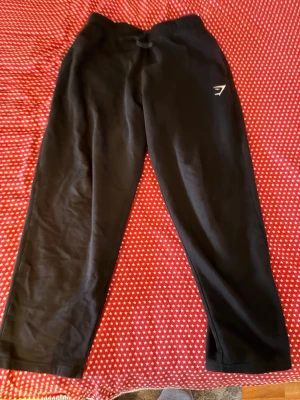  Gymshark - Svarta mjukisbyxor från Gymshark med dragsko i midjan och elastisk resår. Byxorna har en avslappnad passform och är tillverkade i mjukt material, perfekta för chill eller träning. Gymshark-logga i vitt på ena benet.