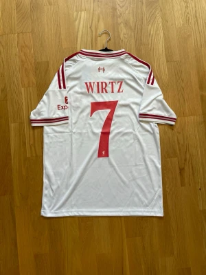 Wirtz #7 - S - Säljer en vit Liverpool matchtröja från Adidas med röda detaljer och trycket WIRTZ 7 på ryggen. Tröjan har V-ringning, röda ränder på axlarna och klubbmärke samt sponsorlogga på bröstet. Perfekt för fotbollsfans som vill sticka ut på planen.