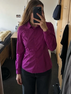 Mango figursydd rosa skjorta – strl S - Snygg skjorta från Mango Basics i en stark cerise/magenta färg. Figursydd modell med markerad midja, klassisk krage och långa ärmar. Detaljer med veck framtill ger en feminin passform.  Storlek S   Fint begagnat skick. Se bilder för passform och skick.