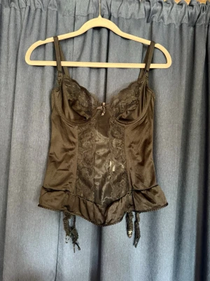 Svart spetsbody med volangkant - Snygg svart body med spetsdetaljer framtill, justerbara axelband och volangkant nedtill. Utrustad med strumpebandshållare och dekorativ rosett mellan kuporna. Materialet är 100% nylon. Budas ut på min live om intresse finns. 