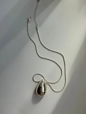 Silverfärgat halsband med droppformad berlock - Snyggt halsband i silverfärg med en stor, droppformad berlock i metall. Kedjan är tunn och smidig med justerbar längd och karbinlås. Perfekt för dig som gillar minimalistisk och modern stil.