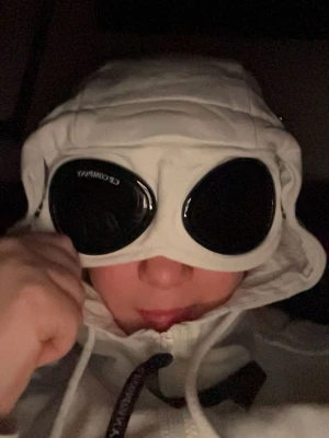 Vit vindjacka med huva och glasögondetalj - Säljer en vit vindjacka med unik huva som har inbyggda glasögon. Jackan har dragkedja framtill och snörning i huvan. Perfekt för dig som vill sticka ut med en futuristisk look.