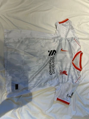 Liverpool match T-shirt  - Snygg vit Liverpool matchtröja från Nike med subtilt mönster i tyget. Tröjan har röda och gula detaljer vid hals och ärmslut, samt klubbmärke och sponsorloggor på bröstet. Perfekt för fotbollsträning eller att bära till vardags.