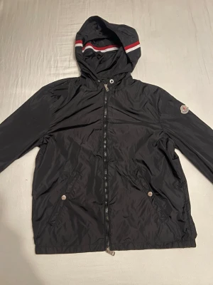 Svart vindjacka från Moncler med huva - Svart vindjacka från Moncler med dragkedja framtill, två fickor med knappar och huva med röd och vit rand. Jackan har Moncler-logga på vänster ärm och är tillverkad i ett lätt, glansigt material som står emot vind. Skriv vid funderingar! Har svar på det mesta!