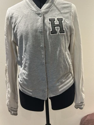Grå bomberjacka/hoodie med H-detalj - Snygg grå bomberjacka/hoodie med vita ärmar och ribbade muddar. Jackan har tryckknappar framtill och en stor H-applikation på bröstet. Den passar mig och jag har storlek S i vanliga fall.