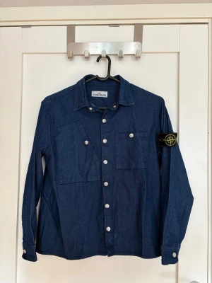 Mörkblå overshirt från Stone Island - Snygg mörkblå overshirt från Stone Island i denimliknande material. Klassisk krage, två bröstfickor och silverfärgade knappar framtill. Ikonisk Stone Island-patch på vänster ärm. Perfekt lager-på-lager-plagg för en cool streetstil.