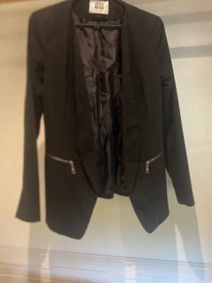 Svart kavaj med dragkedjedetaljer - Stilren svart kavaj från vero Moda med öppen front och två dragkedjorfickor framtill. Kavajen har lång ärm, slät passform och helfoder i blankt tyg. Perfekt för en trendig och modern look.