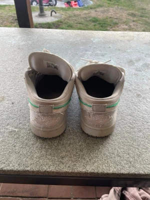 Beige sneakers med grön detalj - Säljer ett par beige sneakers med grön rand på hälen och broderad logga baktill. Skorna har en klassisk låg siluett och är tillverkade i ett material som ser ut som läder. Perfekta för dig som vill ha en stilren och enkel sneaker.