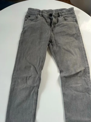 Grå raka jeans - Snygga grå jeans med rak passform och klassisk femficksdesign. Jeansen har bälteshällor, knappgylf och är tillverkade i ett mjukt denimtyg. Perfekta för en avslappnad och stilren look.
