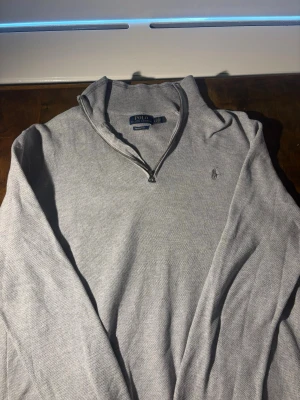 Ljusgrå stickad tröja Polo Ralph Lauren - Ljusgrå stickad tröja från Polo Ralph Lauren med halv dragkedja och klassisk broderad logga på bröstet. Tröjan har lång ärm och ribbade muddar. Perfekt till lager på lager och enkel att matcha med jeans eller chinos.