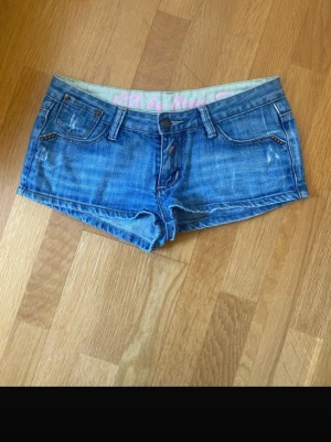 Blå mini denimshorts med nitar - Säljer ett par blå denimshorts med låg midja och snygga nitar på bakfickorna. Shortsen har klassisk femficksdesign, bälteshällor och stängs med knapp och dragkedja. Storlek S/xs Perfekta för sommardagar och festivaler!