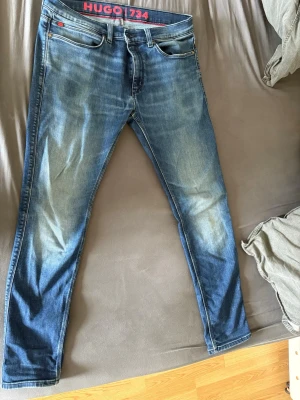 Blå slim fit jeans från HUGO - Snygga blå slim fit jeans från HUGO med klassisk femficksdesign och diskreta slitningar framtill. Jeansen har en något tvättad look och röda detaljer vid linningen. Perfekta för en trendig och avslappnad stil.