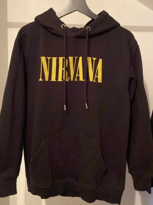 Nirvana hoodie - Svart hoodie med stor gul Nirvana-logga på bröstet och ett stort tryck på ryggen från albumet 'In Utero' med ängelmotiv och text. Hoodien har huva med dragsko och en klassisk magficka. Perfekt för dig som gillar bandmerch och grunge-stil.