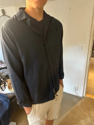 Mörkblå linneskjorta med lång ärm - Snygg mörkblå skjorta i linne med klassisk krage och långa ärmar. Skjortan har knäppning framtill och en avslappnad passform som passar perfekt till både jeans och shorts. Enkel och stilren design utan synliga loggor.