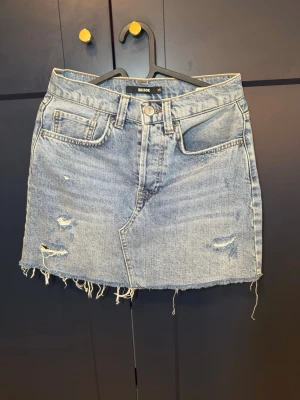 Ljusblå jeanskjol med fransig kant - Säljer en ljusblå jeanskjol från Bik Bok med fransig, rå kant och slitna detaljer framtill. Kjolen har klassisk femficksmodell och knappgylf. Perfekt för en avslappnad och trendig look.passar s också 