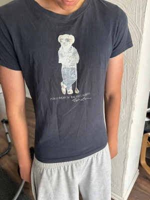 Svart Polo Bear t-shirt från Ralph Lauren - Snygg svart t-shirt från Ralph Lauren med Polo Bear-tryck på bröstet. Klassisk passform och rund halsringning. Tillverkad i mjuk bomull som känns skön mot huden. Perfekt för dig som gillar stilrena och ikoniska plagg.