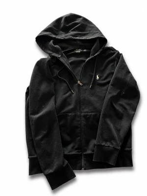 Svart hoodie från Polo Ralph Lauren - Klassisk svart hoodie från Polo Ralph Lauren med dragkedja framtill och justerbar huva. Tröjan har fickor på magen och den ikoniska broderade loggan i vitt på bröstet. Tillverkad i mjuk bomullsblandning för skön komfort.
