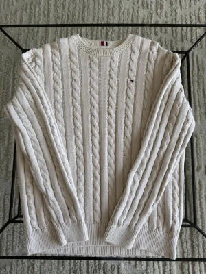 Beige stickad tröja - Kabelstickad tröja i Beige med rund hals och ribbade muddar. Tröjan har ett diskret broderat logomärke på bröstet och klassiska stickade ränder. Perfekt för lager på lager och ger en clean look.