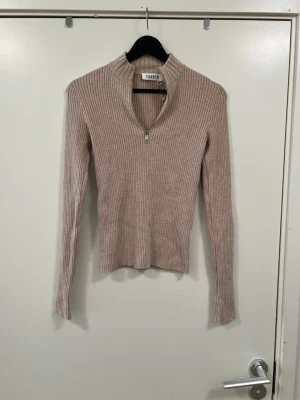 Beige ribbad stickad tröja med zip - Säljer en beige ribbad stickad tröja från EDITED med lång ärm och half zip framtill. Tröjan har en hög krage och är figurnära i modellen, perfekt för lager på lager under höst och vinter.