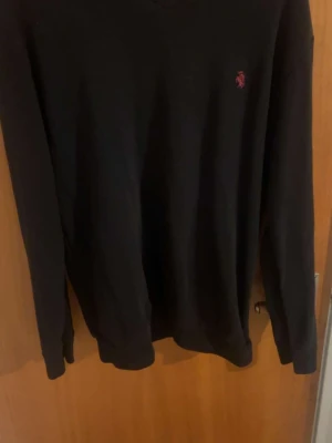 Polo Ralph sweater - Snygg svart stickad tröja från Polo Ralph Lauren med klassisk röd broderad logga på bröstet. Tröjan har V-ringning och långa ärmar med ribbade muddar. Perfekt till lager på lager och enkel att matcha.