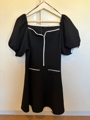 Svart klänning med puffärmar från Zara - ZARA Black & White Dress – New with Tags 🖤🤍  Elegant black mini dress from ZARA with contrasting white trim, puff sleeves and decorative front pockets. Chic and feminine design, perfect for parties, dinners or special occasions.  • Size: L • Color: Black / White • Condition: New with tags • Original price: 379 SEK • Article no.: 2298/459/093  Never worn ✨