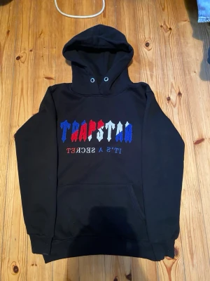 Svart hoodie med TRAPSTAR-brodyr - Svart hoodie med broderad TRAPSTAR-text i blått, rött och vitt på bröstet samt texten 'IT'S A SECRET' under. Klassisk känguruficka och huva med dragsko. Perfekt för en streetwear-look.