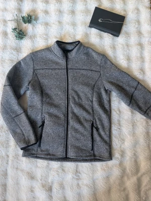 Grå ziptröja med ståkrage - Snygg grå ziptröja med ståkrage och svarta detaljer. Tröjan har hel dragkedja framtill och två fickor med dragkedja. Tillverkad i mjukt, melerat material som passar perfekt till lager-på-lager. Enkel och stilren design.