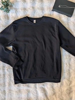 Svart basic tunn sweatshirt - En enkel svart tunn sweatshirt med rund hals och långa ärmar. Tröjan har ribbade muddar vid ärmslut och nederkant, vilket ger en klassisk och clean look. Perfekt att matcha med jeans eller joggers för en avslappnad stil.