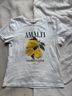 Vit t-shirt med Amalfi citrontryck från HM - Säljer en vit t-shirt med tryck av citroner och texten 'AMALFI VACATION CLUB Italy' på framsidan. T-shirten har rund hals och korta ärmar, perfekt för dig som gillar fräscha och somriga vibes. Materialet känns mjukt och skönt mot huden. S i storlek, lite kortare modell. Aldrig använd. 