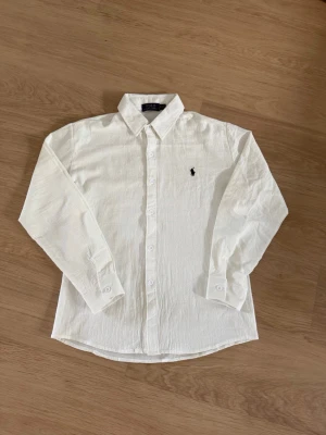 Vit linneskjorta från Polo Ralph Lauren - Klassisk vit skjorta i linne från Polo Ralph Lauren med lång ärm och knappar framtill. Skjortan har en broderad logga på bröstet och en tidlös krage. Perfekt för en clean och stilren look.