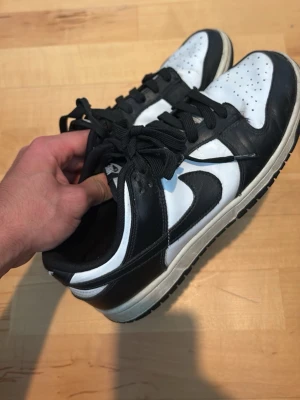 Svarta och vita Nike Dunk Low sneakers - Säljer ett par klassiska Nike Dunk Low sneakers i svart och vitt läder. Skorna har den ikoniska swoosh-loggan på sidan, svart yttersula och vita detaljer på tå och sidor. Lågt skaft och snörning framtill. Perfekta för dig som gillar streetwear och tidlös stil.
