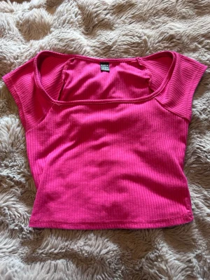 Ribbad rosa crop top från SHEIN - Trendig ribbad crop top i starkt rosa från SHEIN. Toppen har korta ärmar, bred urringning och är gjord i ett stretchigt, mjukt material som sitter snyggt på kroppen. Perfekt att matcha med höga jeans eller kjol för en cool look.