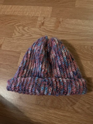Flerfärgad ribbstickad mössa - Mysig ribbstickad mössa i flera färger som rosa, blått och lila. Mössan har uppvikt kant och är gjord i ett mjukt garn som känns skönt mot huden. Perfekt för kalla dagar när du vill ha en färgklick till din outfit.