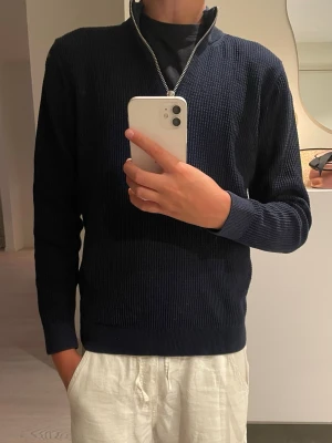 Mörkblå stickad tröja med halvzip - Mörkblå stickad tröja med halvzip och hög krage. Tröjan har lång ärm och ribbade muddar vid ärmslut och nederkant. Perfekt för lager på lager och enkel att matcha till olika outfits.