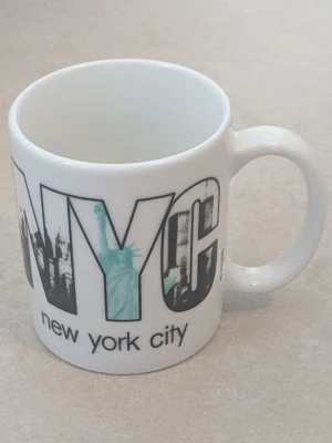 NYC mugg med stadsmotiv - Vit keramikmugg med tryck av texten 'NYC' och motiv från New York City, inklusive Frihetsgudinnan och stadssilhuett i svart och turkos. Under texten står det 'new york city'. Perfekt för kaffe eller te. På undersidan av muggen finns några mörka fläckar, visas på bild ( sista bilden).