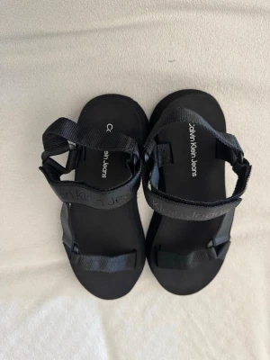 Svarta sandaler Calvin Klein Jeans - Stilrena svarta sandaler från Calvin Klein Jeans med justerbara remmar i textil och logga på ovansidan. Sulan är platt och bekväm, perfekt för sommardagar. Minimalistisk design som passar till många outfits.