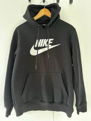 Nike hoodie - Snygg svart hoodie från Nike med stor vit logga och swoosh på bröstet. Klassisk känguruficka framtill, ribbade muddar och justerbar huva med snören. Perfekt för en sportig och avslappnad stil.