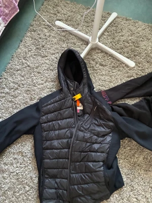 Svart pufferjacka från Parajumpers - Snygg svart pufferjacka från Parajumpers med huva och quiltad front. Jackan har släta ärmar i stretchigt material och dragkedja framtill. Perfekt för kalla dagar och har en cool orange detalj vid halsen. Märkt med 'Warm Up' på insidan.