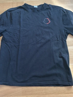 Svart t-shirt med liten logga - En svart t-shirt i bomull med korta ärmar och en liten, diskret logga i rött och vitt på bröstet. Klassisk passform och rund halsringning. Perfekt till jeans eller shorts för en enkel och clean look.