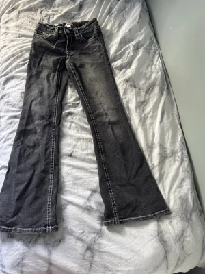 Svarta bootcut jeans med kontrastsömmar - Säljer ett par svarta bootcut jeans med vita kontrastsömmar och snygga detaljer på bakfickan. Jeansen har hög midja och utsvängda ben, vilket ger en retro vibe. Perfekta för dig som vill sticka ut lite extra.