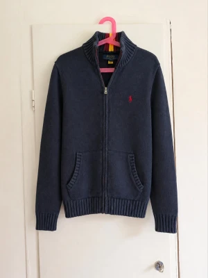 💙 Polo Ralph Lauren stickad zip-kofta – M (10–12) - Klassisk marinblå stickad tröja/kofta från Polo Ralph Lauren med hel dragkedja. Röd broderad ponny på bröstet och snygga röd/gula detaljer i den ribbade kragen. • Storlek: M (10–12 år) • Färg: Marinblå • Hel dragkedja • Två fickor framtill • Broderad röd Polo-logga • QR-tagg finns kvar • Fint begagnat skick men lite färg blekt! Väldigt snygg och tidlös modell som passar perfekt till preppy/Stockholm/grisch-stil. Skriv gärna vid frågor eller för mått!