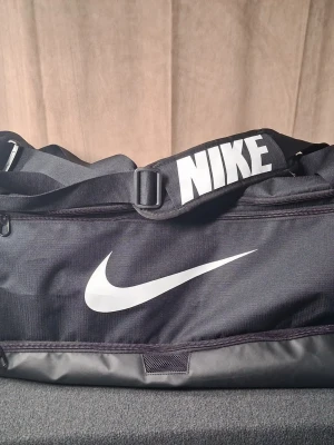 Svart sportbag från Nike - Snygg svart sportbag från Nike med stort vitt logotryck på sidan och justerbar axelrem med vaddering. Väskan har flera dragkedjefack och är tillverkad i slitstarkt syntetmaterial. Perfekt för gymmet eller resan när du vill ha plats för allt.