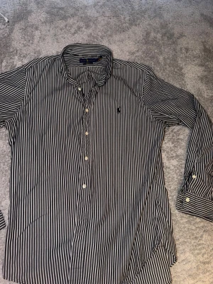 NY Randig skjorta från Ralph Lauren - Säljer en svart och vit randig skjorta från Ralph Lauren i slim fit-modell. Skjortan är tillverkad i stretchig bomull och har klassisk krage, långa ärmar och knappar framtill. Perfekt för dig som gillar stilrena och tidlösa plagg.