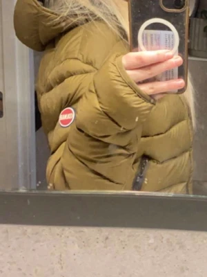 Olivgrön dunjacka med huva - Snygg brun dunjacka med huva och svarta dragkedjor. Jackan har ett quiltat mönster och en rund röd-vit patch på ärmen. Perfekt för kalla vinterdagar och har två praktiska fickor med dragkedja.