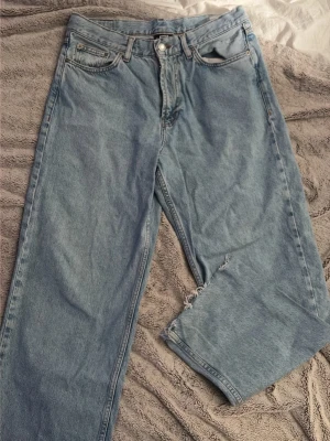 Ljusblå raka jeans med slitning - Säljer ett par ljusblå jeans med rak passform. Jeansen har en tydlig slitning och reva på benen för en trendig, avslappnad look. Tillverkade i denim med dragkedjegylf och knapp. Märket är Sweet SKTBS men köpta på Junkyard.